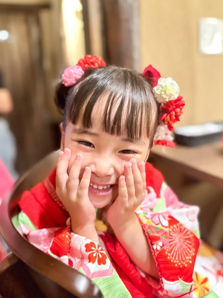 A 3-year-old girl in a Shichi-Go-San kimono smiles with both hands on her cheeks.