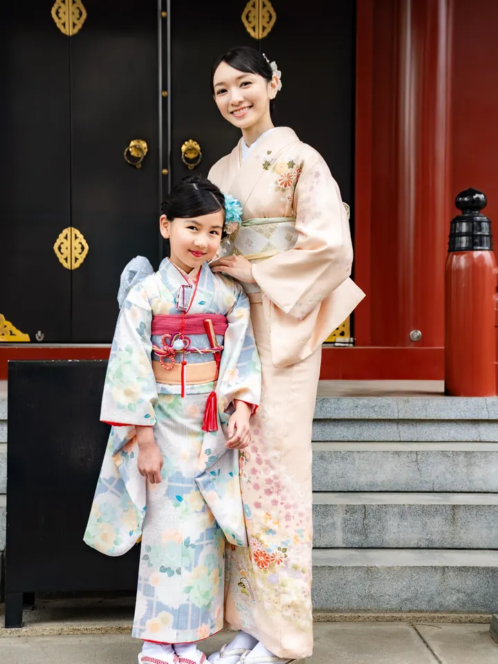 A 7-year-old girl in a kimono stands next to her mother in front of a shrine.